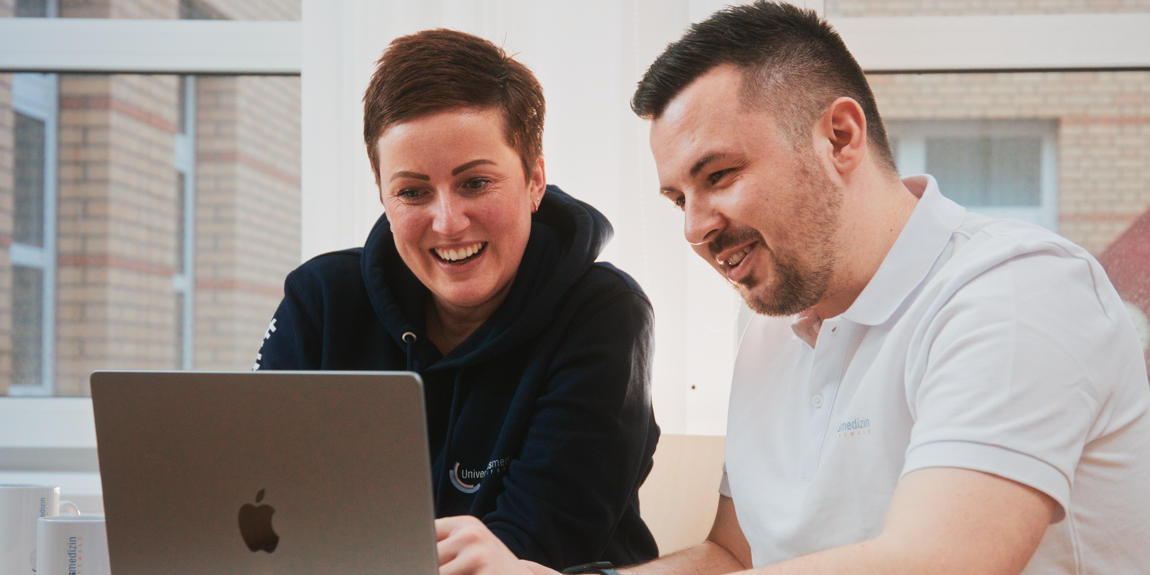 Mitarbeiter der Universitätsmedizin Greifswald Zwei UMG-Mitarbeiter teilen einen heiteren Moment während der Arbeit an einem gemeinsamen Projekt.